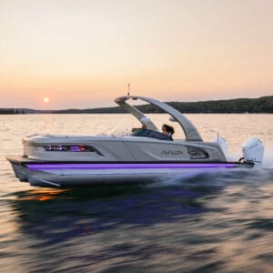 A modern speedboat named Excalibur Elite Windshield cruises swiftly across a calm lake during sunset. The boat's sleek design is highlighted by a subtle neon light strip along its side. A person is seated at the helm, steering the boat. Scenic shoreline and forested hills are in the background.