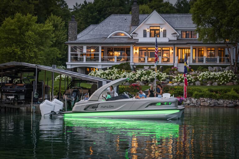 A luxury lakeside house with large windows and a wrap-around porch is illuminated in the evening. In front, an Excalibur Elite Windshield with green LED lights glides on the calm water. People are visible on the boat and dock, surrounded by lush greenery and flags.