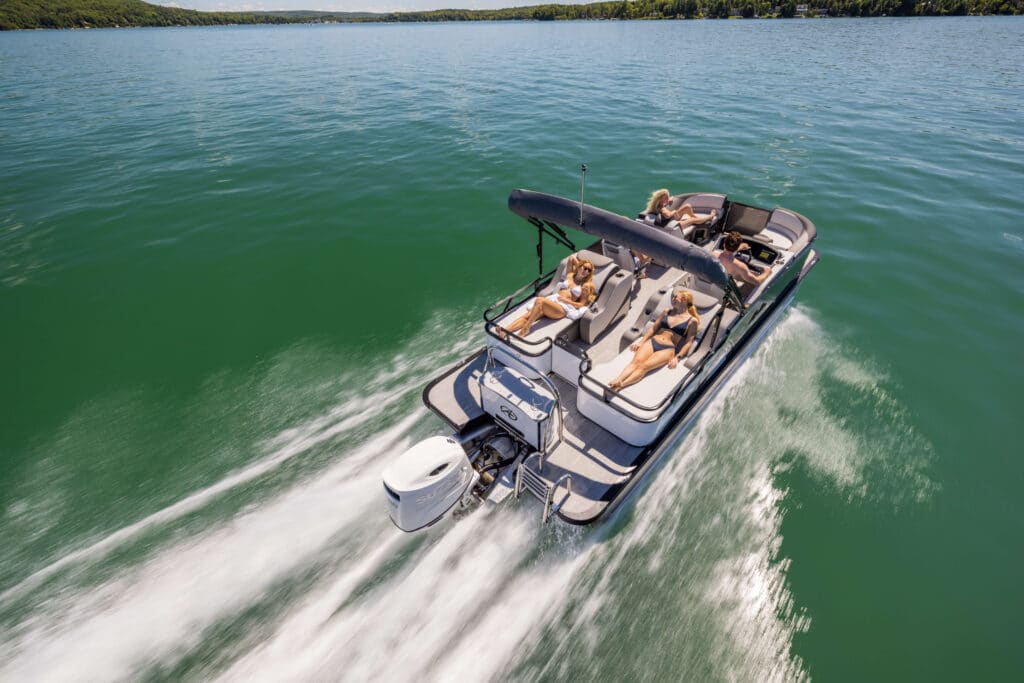 A motorboat with an outboard engine cruises on a clear, greenish-blue lake. A canopy shades the seating area where multiple people are lounging and enjoying the sunlight. The LSZ Rear Lounger leaves a trail of white wake as it moves forward. Trees line the distant shore.