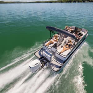 A motorboat with an outboard engine cruises on a clear, greenish-blue lake. A canopy shades the seating area where multiple people are lounging and enjoying the sunlight. The LSZ Rear Lounger leaves a trail of white wake as it moves forward. Trees line the distant shore.