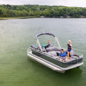 A group of three people is on a VTX Quad Fish in a serene lake. One person is standing and fishing, while the others are seated, enjoying the view. The backdrop features dense, green trees along the shoreline and calm waters reflecting the sky.