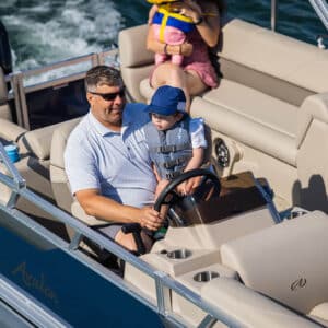 A man and a baby wearing life jackets are seated at the helm of a VLS Quad Lounge with relaxed seating. The baby is sitting on the man's lap, holding the steering wheel. Another adult, holding a child, sits in the background on a bench seat. The boat is on a body of water.
