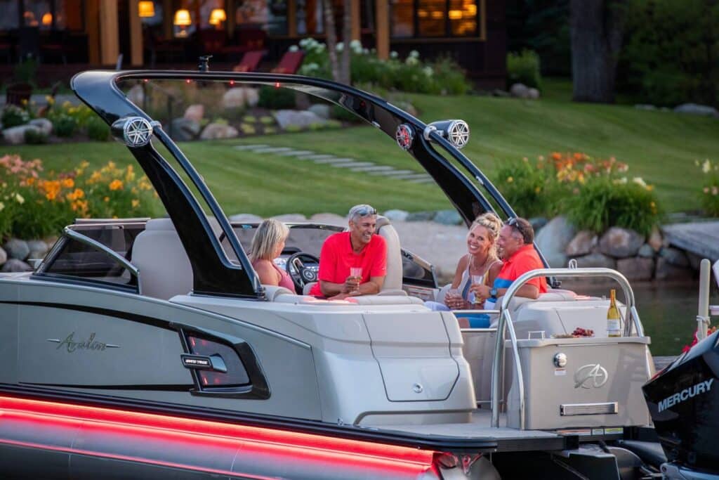 A group of people are sitting and talking on a modern pontoon boat with a silver exterior, docked by a lush garden with flowers. The boat features sleek black railings, and an open area with comfortable seating. A bottle of wine and glasses are placed on a table.