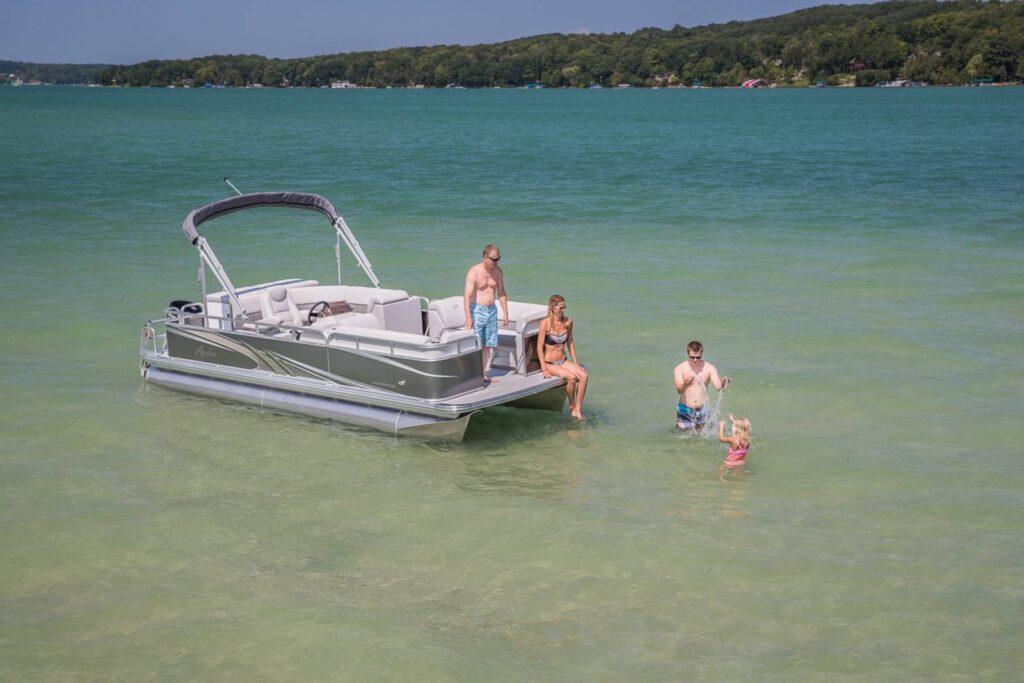 Four people enjoy a sunny day on a pontoon boat in shallow, clear green water near a lush, tree-lined shore. One person is on the boat while the others are in the water, with one person holding a young child.