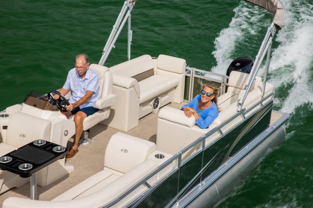 An elderly man steers a pontoon boat with a canopy on a lake, while an elderly woman relaxes on the cushioned seating, smiling and wearing sunglasses. The boat speeds through the water, leaving a trail of white foam behind.