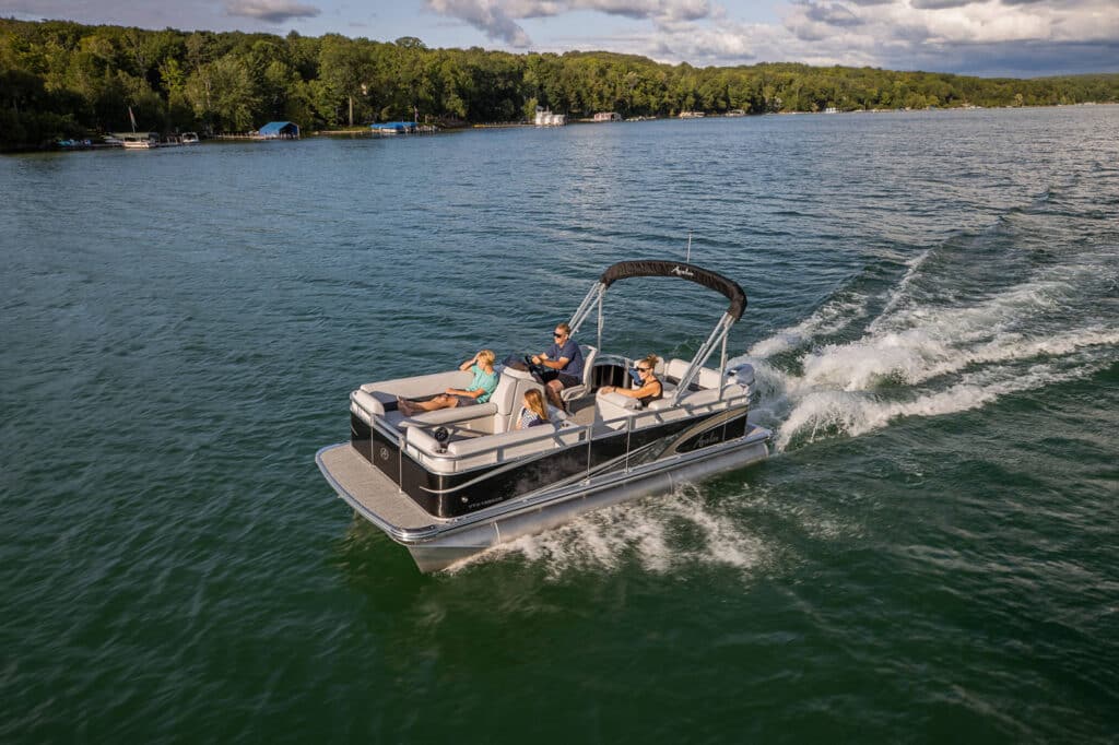 A pontoon boat with five people is cruising on a lake with lush green trees lining the distant shore. The boat leaves a small wake behind as it moves through the calm water under a partly cloudy sky. Some passengers are relaxing on deck seats.