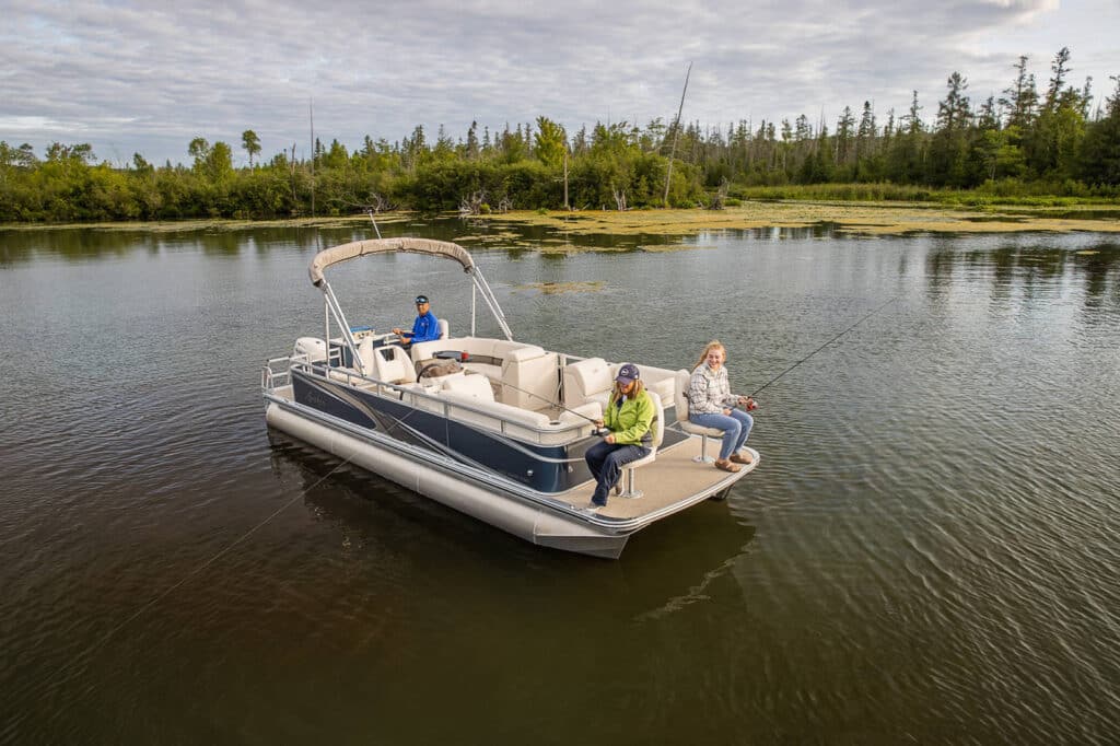 Three people are fishing from a pontoon boat on a calm lake surrounded by trees and greenery. The sky is partly cloudy. One person is seated at the front, another stands near the middle, and the third person is seated near the rear.
