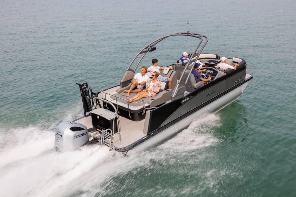 A pontoon boat speeds through calm waters with six people aboard. The passengers are seated comfortably, enjoying the ride, while the boat driver navigates. The boat features a Honda outboard motor, plush seating, and an arching canopy.