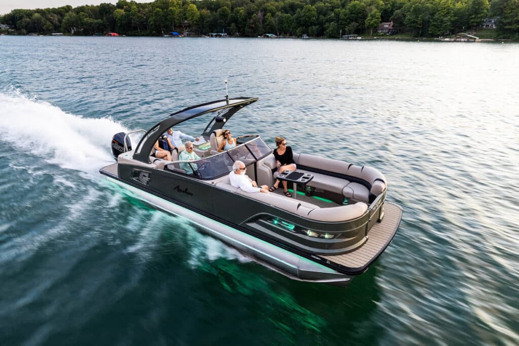 A sleek motorboat glides across a calm lake with five people onboard enjoying the ride. The boat has a modern design with a spacious deck and comfortable seating. The water is clear and serene, with a lush, green treeline in the background.