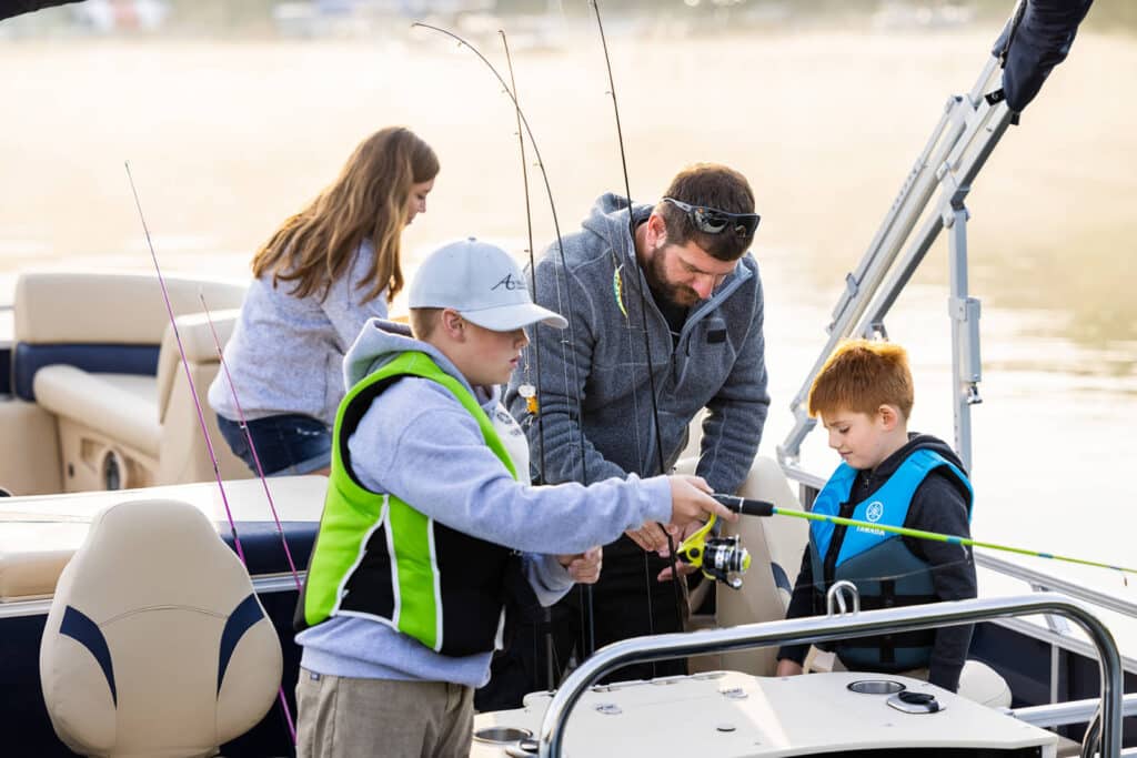 A family is on a boat during a fishing trip. The father assists the young son while holding a fishing rod. The older boy is fishing, and the daughter is in the background. Everyone is dressed in casual clothing, and the children have life jackets on.