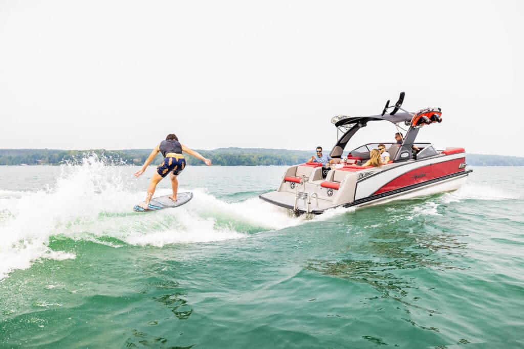 A person wakeboarding on a lake while being towed by a red and white boat. Several people are on the boat, watching the wakeboarder. The lake is calm, with distant green trees visible on the horizon. The sky is clear and sunny.