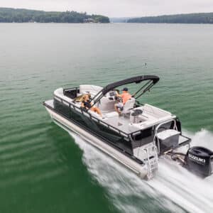 A pontoon boat with a black canopy speeds across a greenish lake. Three people are on board: one steering, one sitting beside the driver, and one lounging at the bow. Forested hills surround the lake under a cloudy sky. A "Venture 85 Elite Windshield" is attached to the boat.