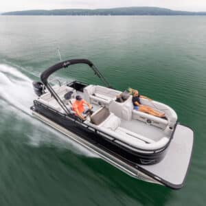 A Venture 85 Cruise with a black canopy is cruising on a calm lake. One man in an orange shirt is steering the boat, while a woman in blue attire is relaxing on the back seat. The surrounding area features serene, forested shoreline under a cloudy sky.