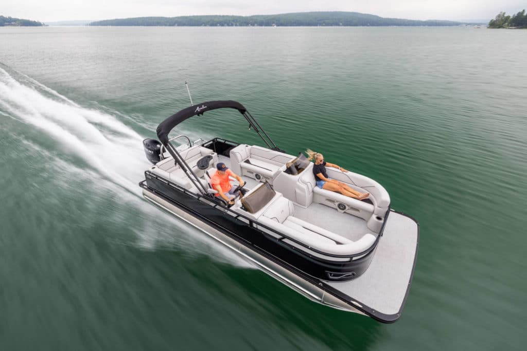 A Venture 85 Cruise with a black canopy is cruising on a calm lake. One man in an orange shirt is steering the boat, while a woman in blue attire is relaxing on the back seat. The surrounding area features serene, forested shoreline under a cloudy sky.