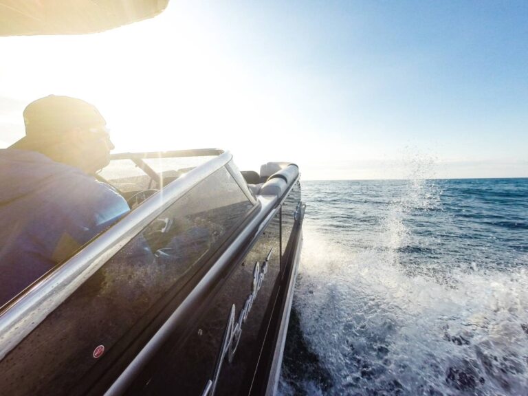 A person in a blue jacket and baseball cap steers a speeding boat over a sunlit ocean. Water splashes up dramatically on the side of the boat. The sky is clear and blue, indicating a bright and sunny day.