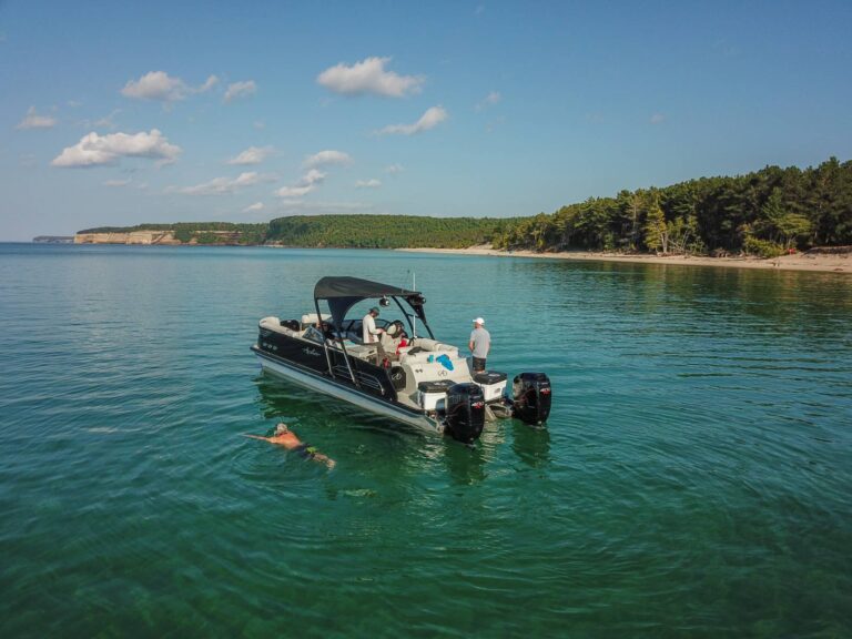 A person swims near a white motorboat with a black canopy floating in clear, calm waters near a tree-lined shoreline under a sky with scattered clouds. Green hills and cliffs are visible in the background.