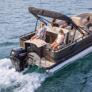 A Venture 85 Rear Fish cruising on a lake, with four people on board. Two children are seated at the back, while an adult is driving and another adult sits nearby. The boat has a black canopy, a Mercury outboard motor, and calm water surrounding it.