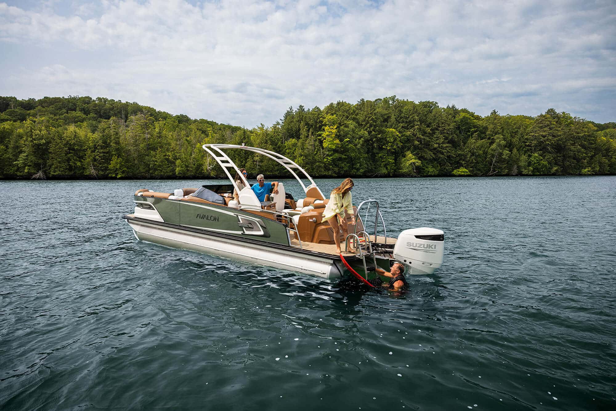 A group of people enjoy leisure time on a silver Excalibur LTD Elite Windshield with a Suzuki outboard motor. One person is swimming near the boat, while another is about to enter the water. Lush green forests line the lake in the background under a cloudy sky.