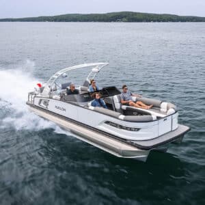 An Excalibur LTD Quad Lounge Windshield speeds across a calm lake with four people onboard. The boat has a sleek, modern design, and the passengers are seated comfortably, enjoying the ride. The background features a tree-lined shoreline under a clear sky.
