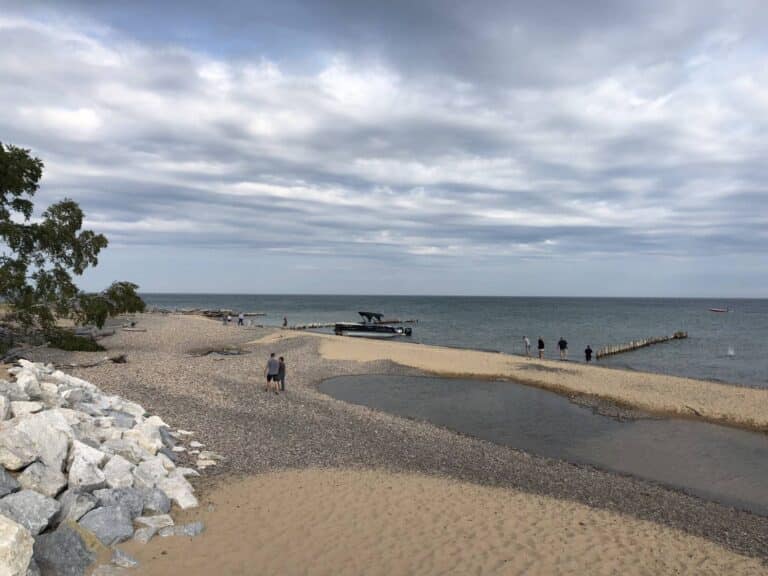 A pebbled and sandy beach by the sea under a cloudy sky. Two people walk along the shore, a small motorboat is anchored close to the coast, and several people can be seen near the water. Rocky embankment and sparse vegetation are present on the left.