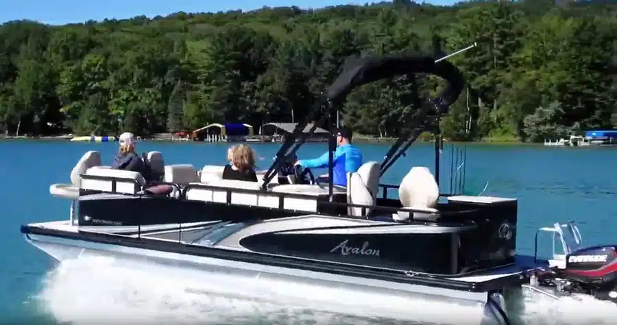 An avalon pontoon boat with three passengers riding in front of the lake shoreline.