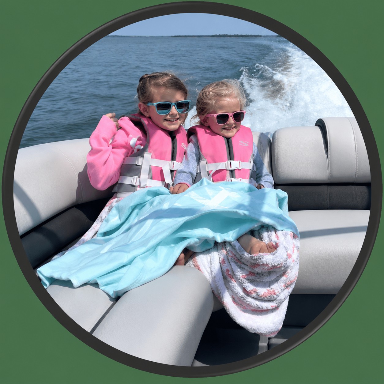 Two young girls wearing pink life jackets and sunglasses sit on a boat, wrapped in blankets. They smile and sit close together as the boat rides on a sunny day, with water and waves visible in the background.