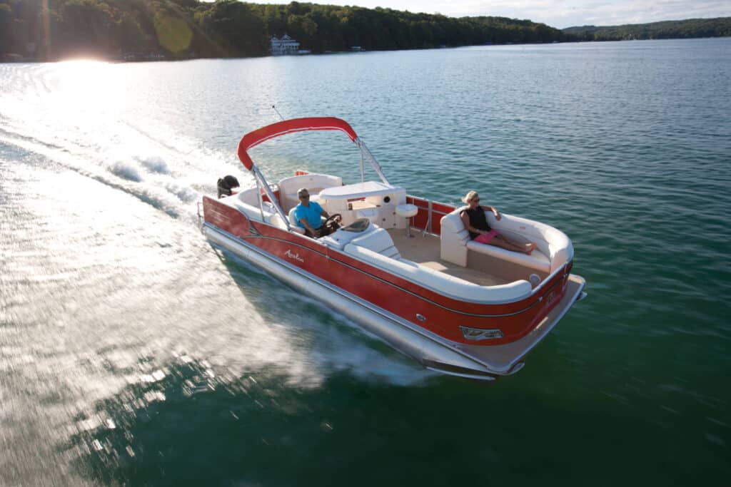 A red and white pontoon boat cruises on a large body of water with a forested shoreline in the background. Two people are on the boat; one person is steering at the helm while the other is reclining on a seat at the back, enjoying the sunny day.