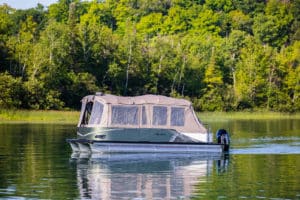 A pontoon boat with a canopy floats on a calm lake. The boat is partially covered, providing shade and shelter. Surrounding the lake, dense, lush green trees reflect beautifully on the water's surface under a clear sky.