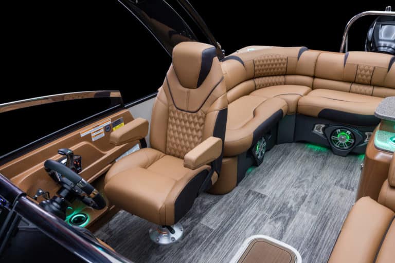 A luxurious boat interior featuring tan leather seats with diamond stitching, including a captain's chair next to the steering wheel, and a curved seating area. The floor has a wood-like appearance, and there are green LED accent lights under the seats.