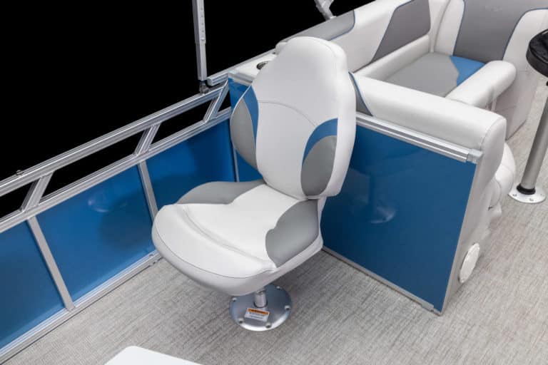 A white captain's chair with gray and blue accents is mounted on a pontoon boat's deck. The chair is located near the side railing, which has blue paneling. Behind the captain's chair, part of a cushioned bench seat is visible. The flooring is light-colored.