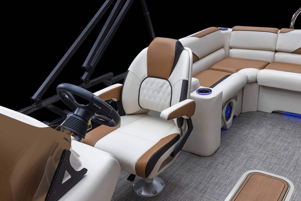 A close-up of the driver's seat on a luxury pontoon boat. The seat is upholstered in white and brown leather and is positioned behind a steering wheel with controls. The spacious interior seating area is visible in the background, featuring similar upholstery.