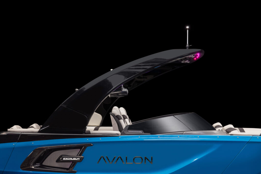 A close-up side view of a modern blue Avalon boat with white seats, a black arch, and a small antenna, set against a solid black background.