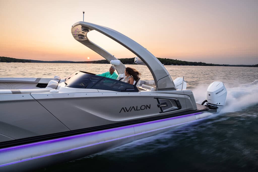 A sleek, white motorboat labeled "Avalon" speeds across a body of water at sunset. Two people are seated on the boat, enjoying the ride. The sun is low on the horizon, casting a warm glow across the water and sky. The boat's trim features a subtle purple light.