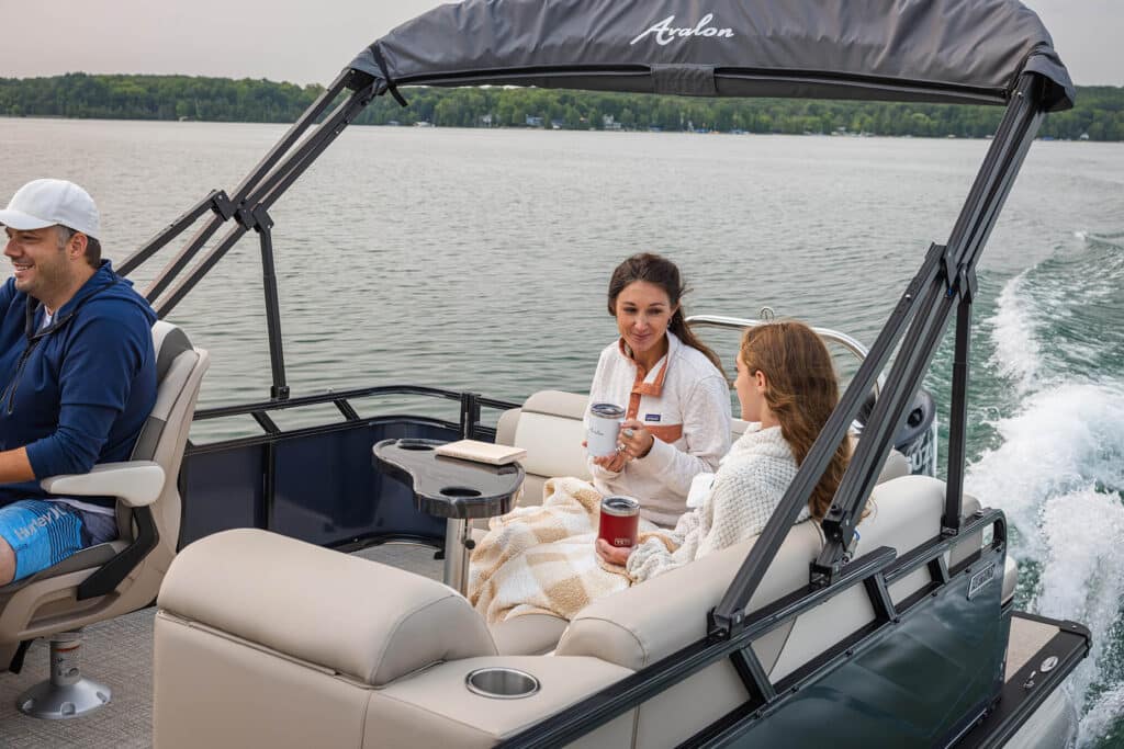 Three people are on a boat with one person seated at the helm and two women sitting on a cushioned bench facing each other, holding mugs. The boat is moving through calm water towards a green, tree-lined shore. The women are covered with a blanket.