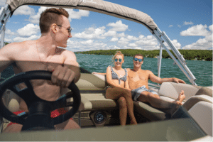 Three individuals enjoy a sunny day on a boat. A man in sunglasses and swim trunks steers the boat, while a woman in a swimsuit and sunglasses sits next to another man in sunglasses and swim trunks. They are all smiling, with a green shoreline and a blue sky in the background.