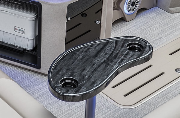 An image of a kidney-shaped, black, marbled table with two built-in cup holders, mounted on a pedestal in a boat interior. The table is situated next to what appears to be a storage compartment and a built-in speaker, with the floor covered in a light-colored vinyl.