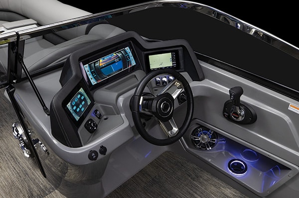 The image shows a modern boat console with multiple display screens, a joystick control, various buttons and switches, a steering wheel, and illuminated cup holders. The dashboard design is sleek with metallic and black elements.