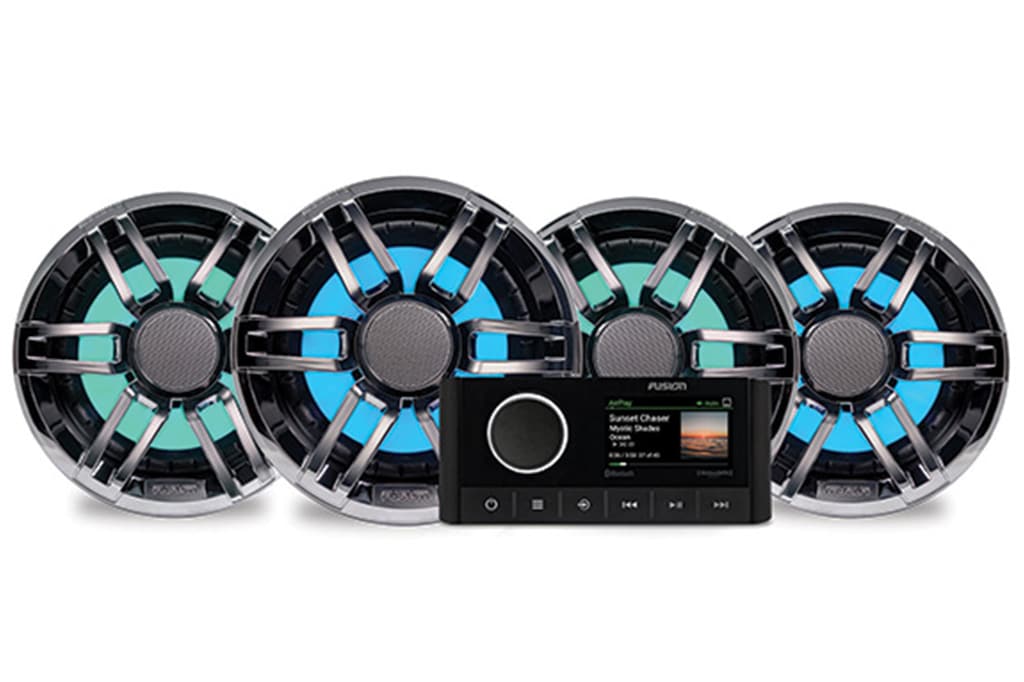 A set of four circular boat speakers with blue and green LED lighting, aligned in a row. In front of the speakers, there is a black marine stereo unit with a display screen showing song information, various buttons, and a control dial.