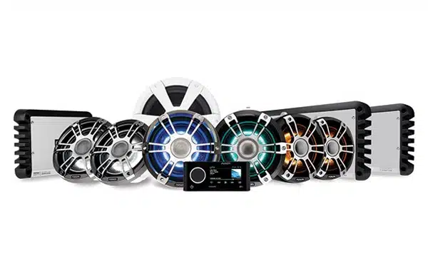 A collection of marine audio equipment displayed on a white background. The setup includes multiple circular marine speakers with colorful LED lights, two amplifiers on either side, and a central marine stereo receiver.