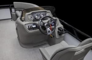 The image shows the driver's console and seating area of a pontoon boat. The console is equipped with a steering wheel, gauges, switches, and various controls. A cushioned chair is positioned in front of the console. The floor is covered with gray carpeting.