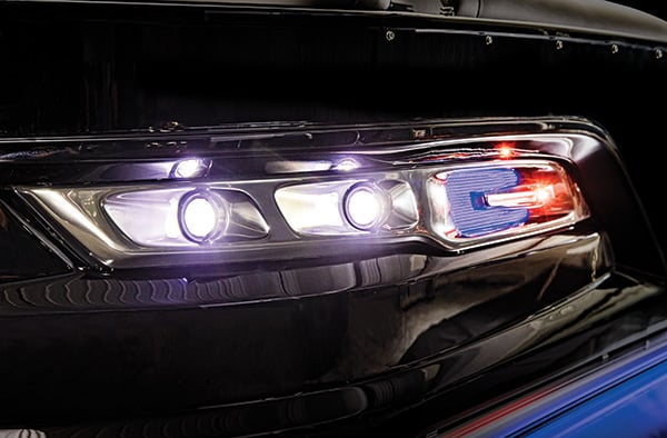 Close-up image of the front headlights of a vehicle, including two bright white lights and a section with red and blue lights. The shiny black surface of the car reflects the lights, giving it a polished look.