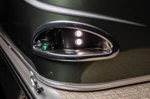 Close-up of the rear left corner of a motorboat. The green and silver boat features an oval-shaped tail light with two white lights and one green light. The edge of the boat is lined with a metal rail, and the flooring appears to be textured fabric or carpet.