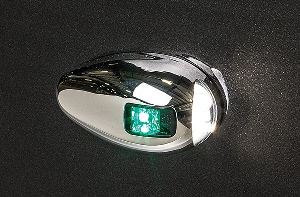 Close-up view of a shiny, oval-shaped, metallic object with a small green light illuminated in its center, against a dark background. The object reflects its surroundings, revealing some bright spots and structures.