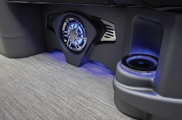 Close-up of a gray car interior featuring a decorative round metallic vent with blue LED lighting, gray upholstery, and a built-in cup holder with soft blue lights underneath. The floor is covered in light gray carpet.