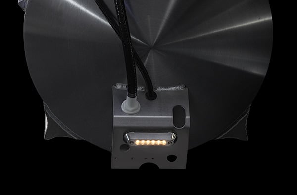 A close-up image of the bottom side of a metallic device with a brushed finish. Wires are extending from the top, and a small rectangular component with multiple LED lights is attached to the bottom, emitting bright light against a dark background.