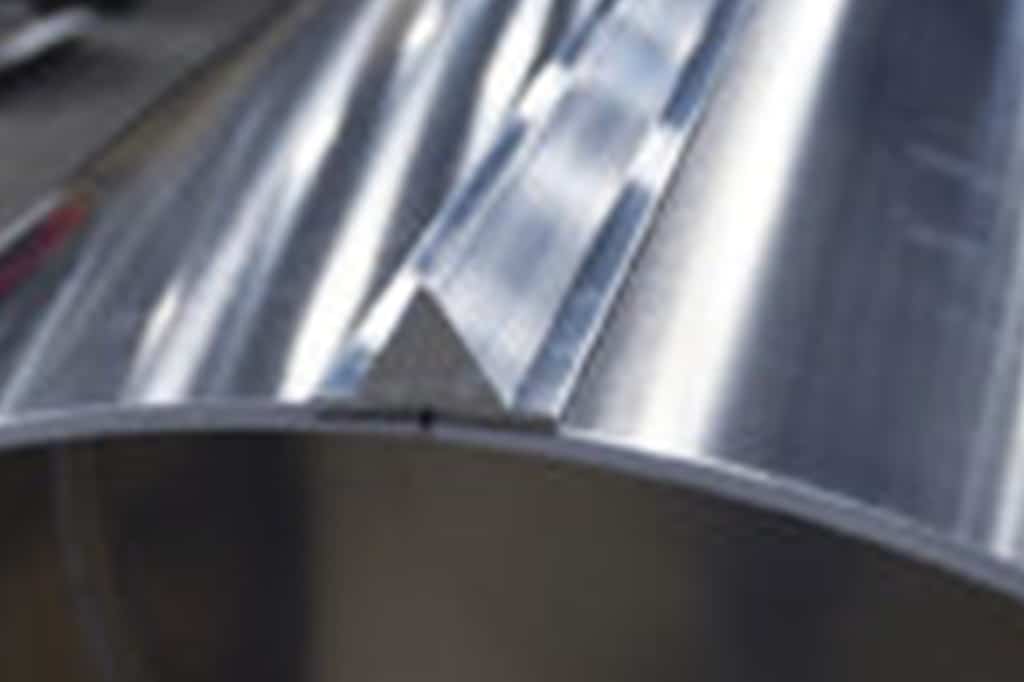 Close-up shot of a shiny, metallic, curved surface with a distinct seam running through the middle. The surface appears reflective, with light creating patterns and shadows across it, highlighting the smooth texture of the metal.