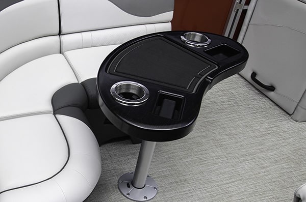 Interior of a boat showing a curved, black table with two cup holders, a smaller square slot, and a flat surface. The seating area behind is upholstered in white and gray. The floor is carpeted with a light gray texture.