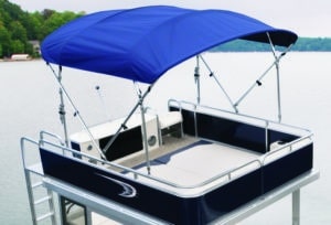 A pontoon boat with a blue canvas shade cover is docked by the water. The boat has a railing around the deck and is equipped with a console and seating area. Trees and a shoreline are visible in the background.