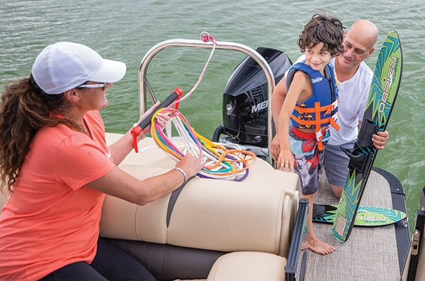 A woman in an orange shirt passes colorful ropes to a man holding a child. They are on a boat with an outboard motor near water. The child, wearing a life vest and holding a wakeboard, appears excited and ready for water sports.