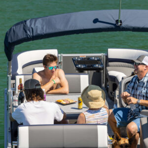 Four people are sitting and chatting on a boat. They are enjoying snacks and drinks on a table in front of them. The boat is on calm, green water. A dog is partially visible near their feet. The atmosphere is relaxed and sunny.